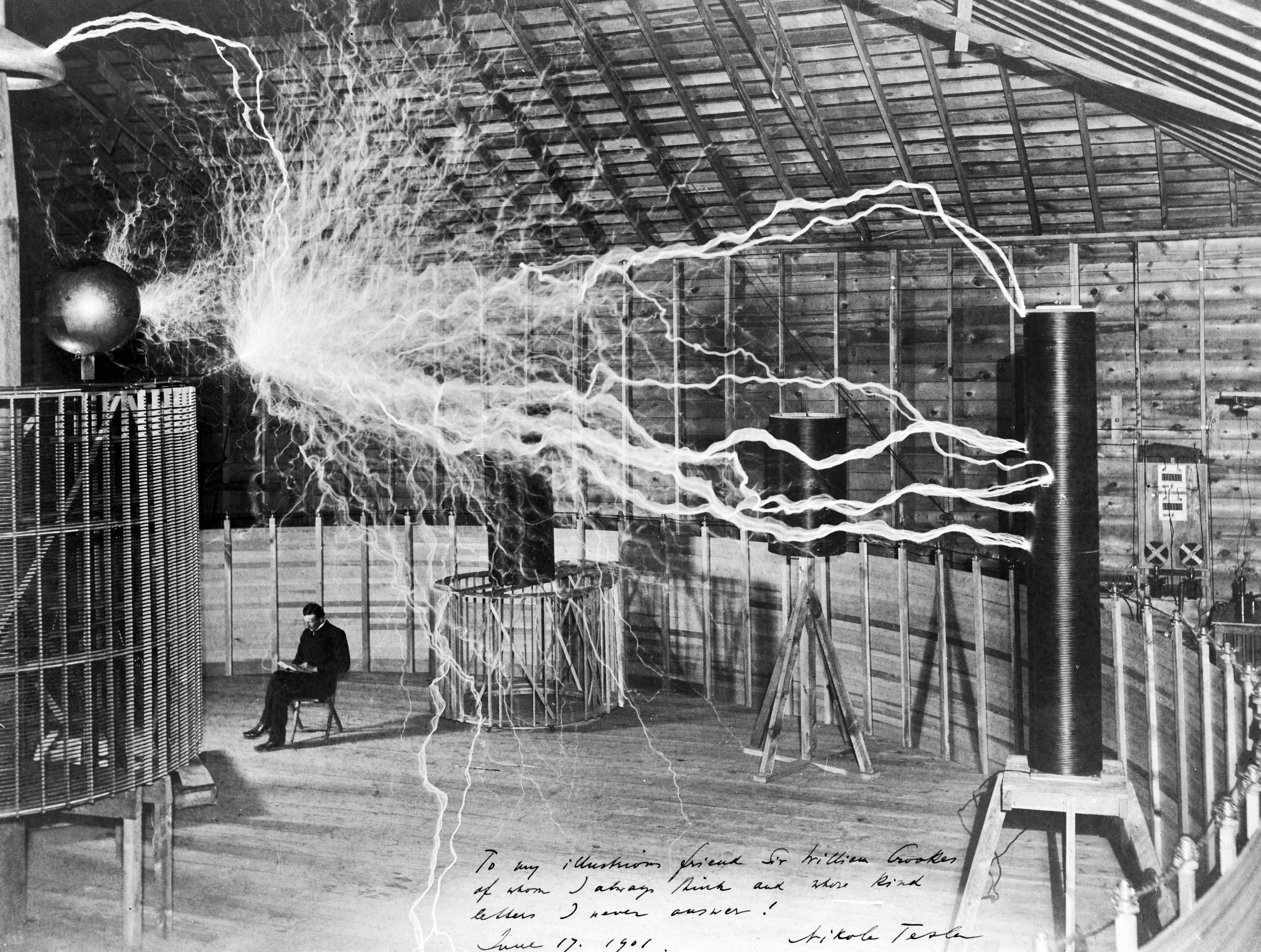 Famous photograph of Serbian-American inventor Nikola Tesla in his laboratory in Colorado Springs around 1899, supposedly sitting reading next to his giant “magnifying transmitter” high voltage generator while the machine produced huge bolts of electricity, Source: Wikipedia