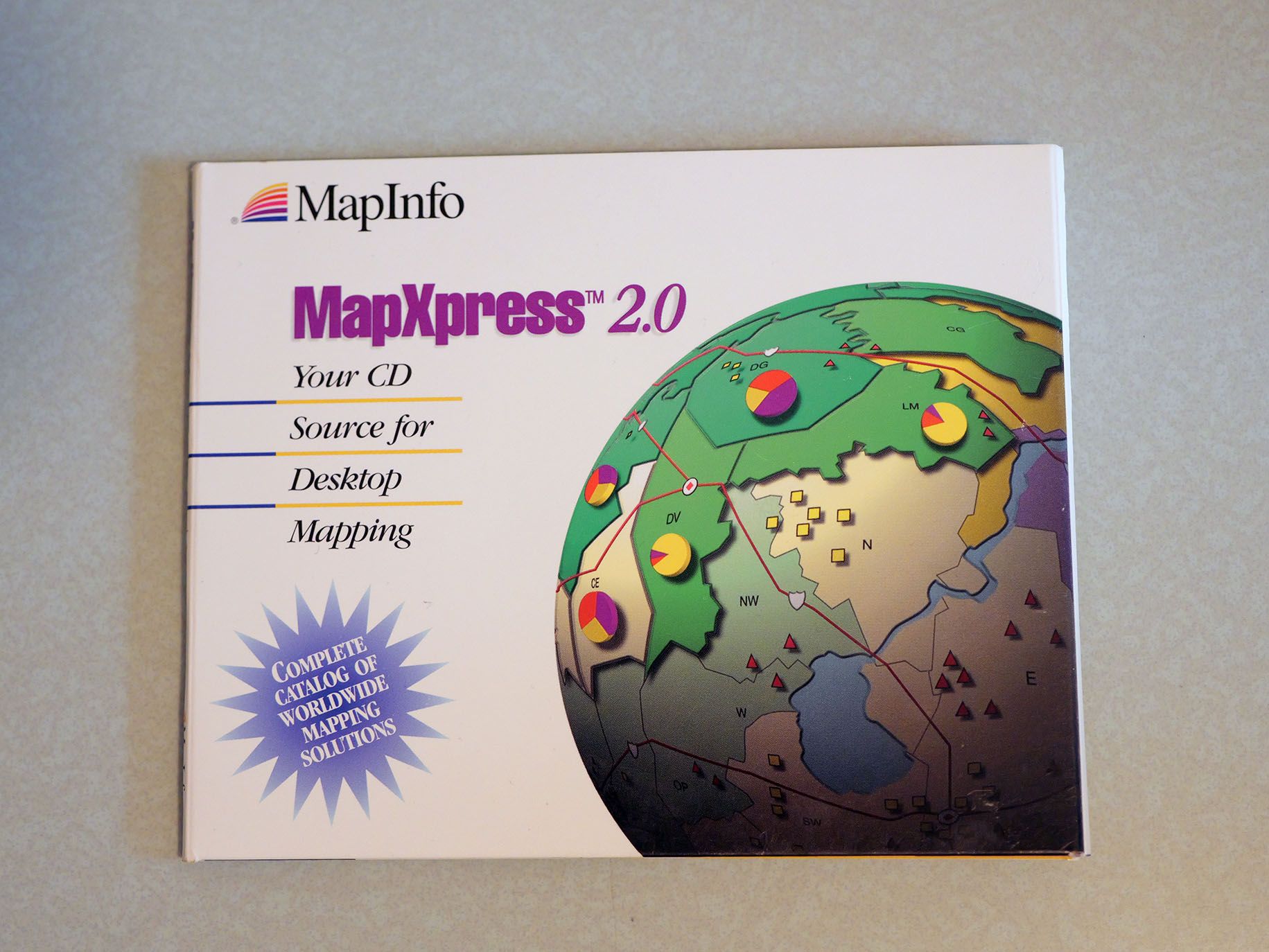 the CD case for MapXpress 2.0, which has the taglines 'Your CD Source for Desktop Mapping' and 'Complete catalog of worldwide mapping solutions.' There's a sphere on this one too, but instead of the graticule logo it's a stylized world map of green land masses and water bodies in polygons, with pie charts on some of the polygons that are sort of raised on the sphere? I am a little skeptical that MapInfo could actually design a map that looked like this.