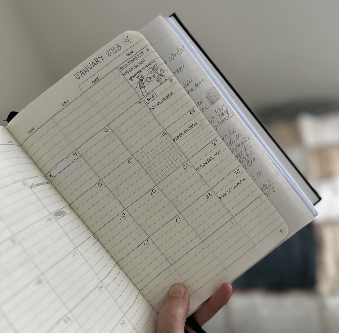 An open A5 Moleskine Cahier notebook being held up at an angle, showing a roughly hand drawn month spreading over both pages