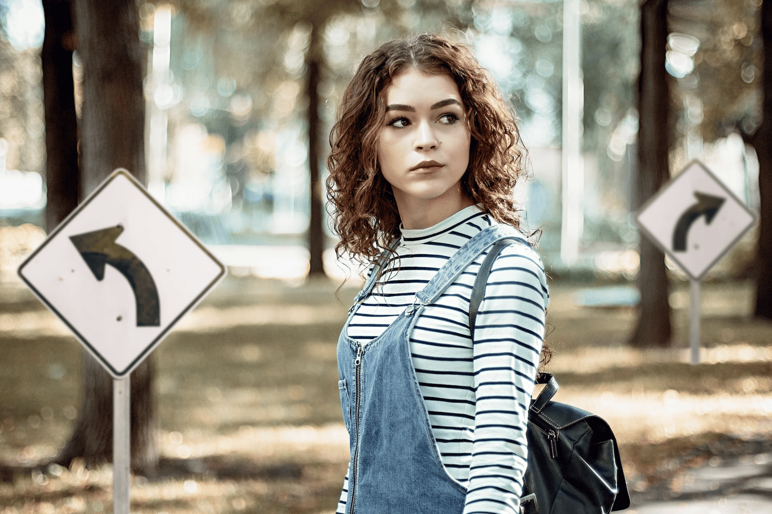 Side view of a girl in a forest facing looking back the way she’s come. There are arrow signs in front and behind her.