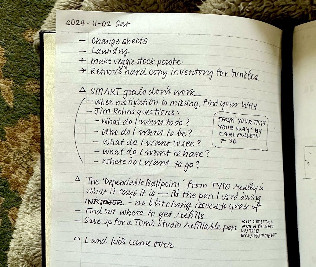 Handwriting in black ballpoint pen on a white page in a notebook that is sitting on a blanket, with a carpet in the background. The page shows a series of notes classified using the Dash-Plus system.