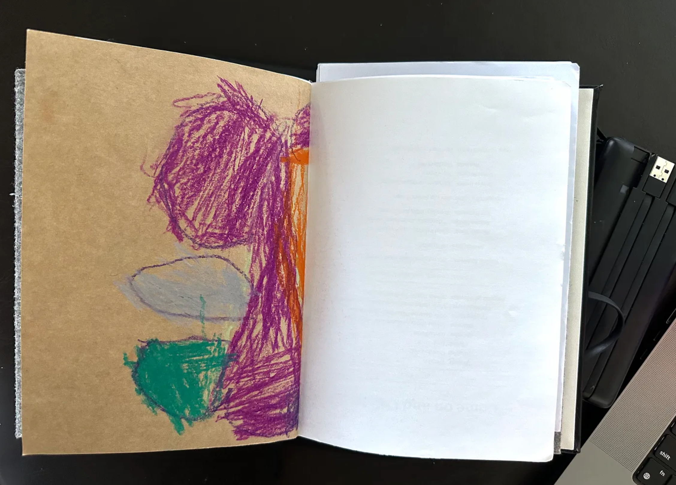 An open notebook on a dark background, with a child’s crayon drawing on brown paper on the left hand page.