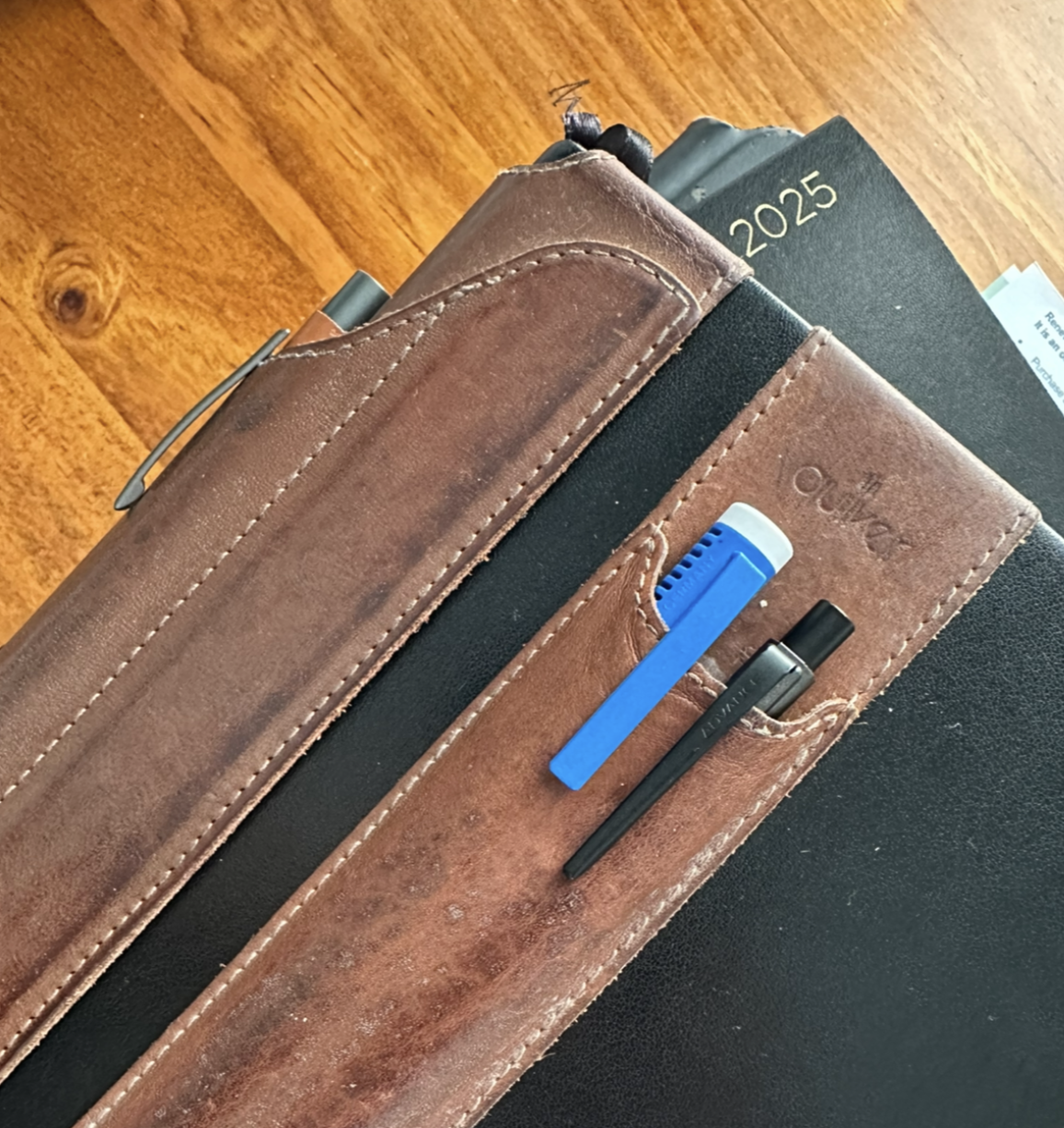 Black notebook, closed, with two tan leather pen holders strapped to the outside. there’s a bright blue eraser pen with a white cap and a mechanical pencil in the double pen holder on the front of the book, and a tan coloured clicky ballpoint pen in the holder that cradles the spine.