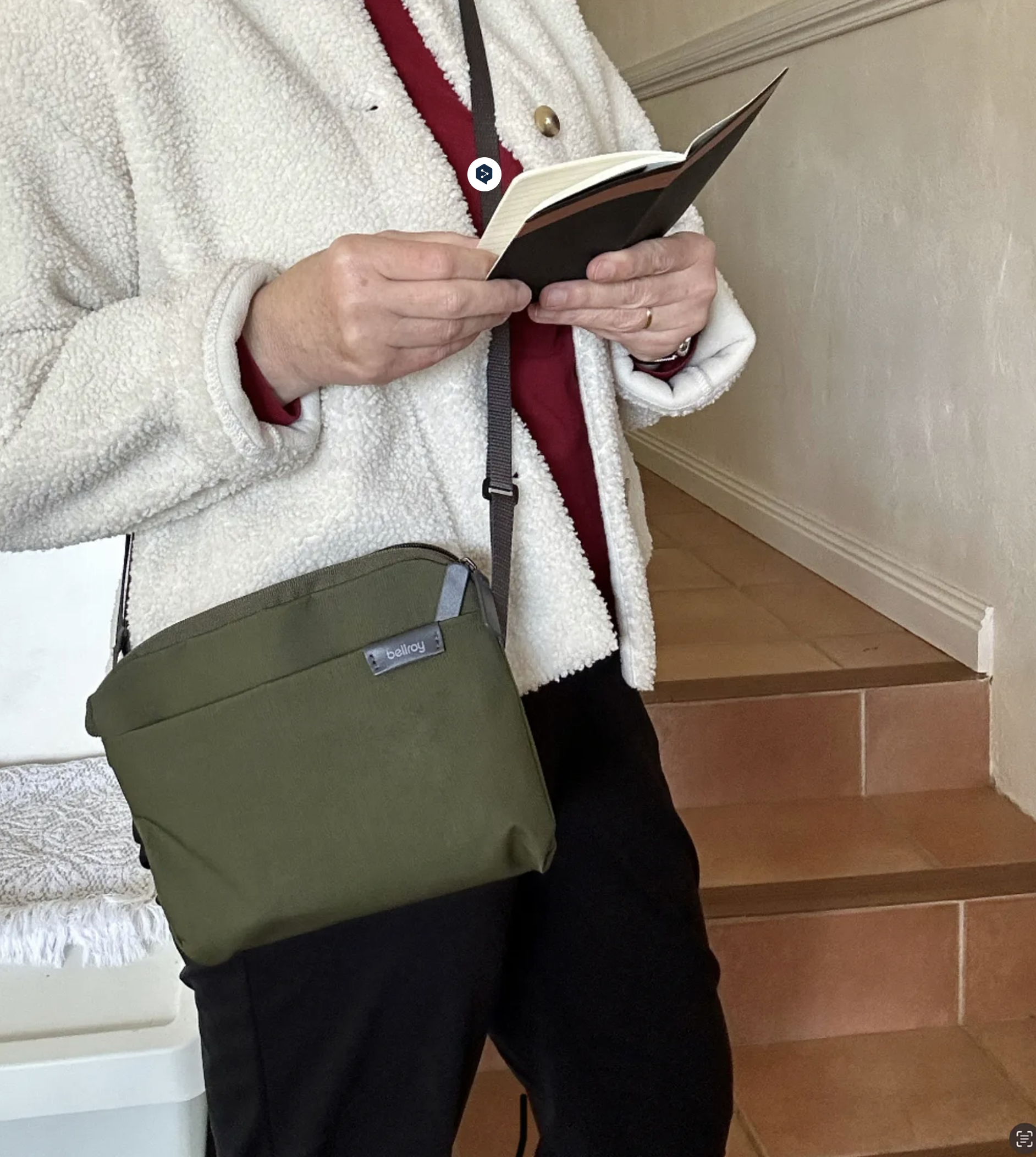 Torso of a person wearing black trousers and a fluffy cream jacket, standing in front of some stairs. They are wearing a khaki-coloured, compact cross body bag, and holding a opened, pocket sized black notebook.