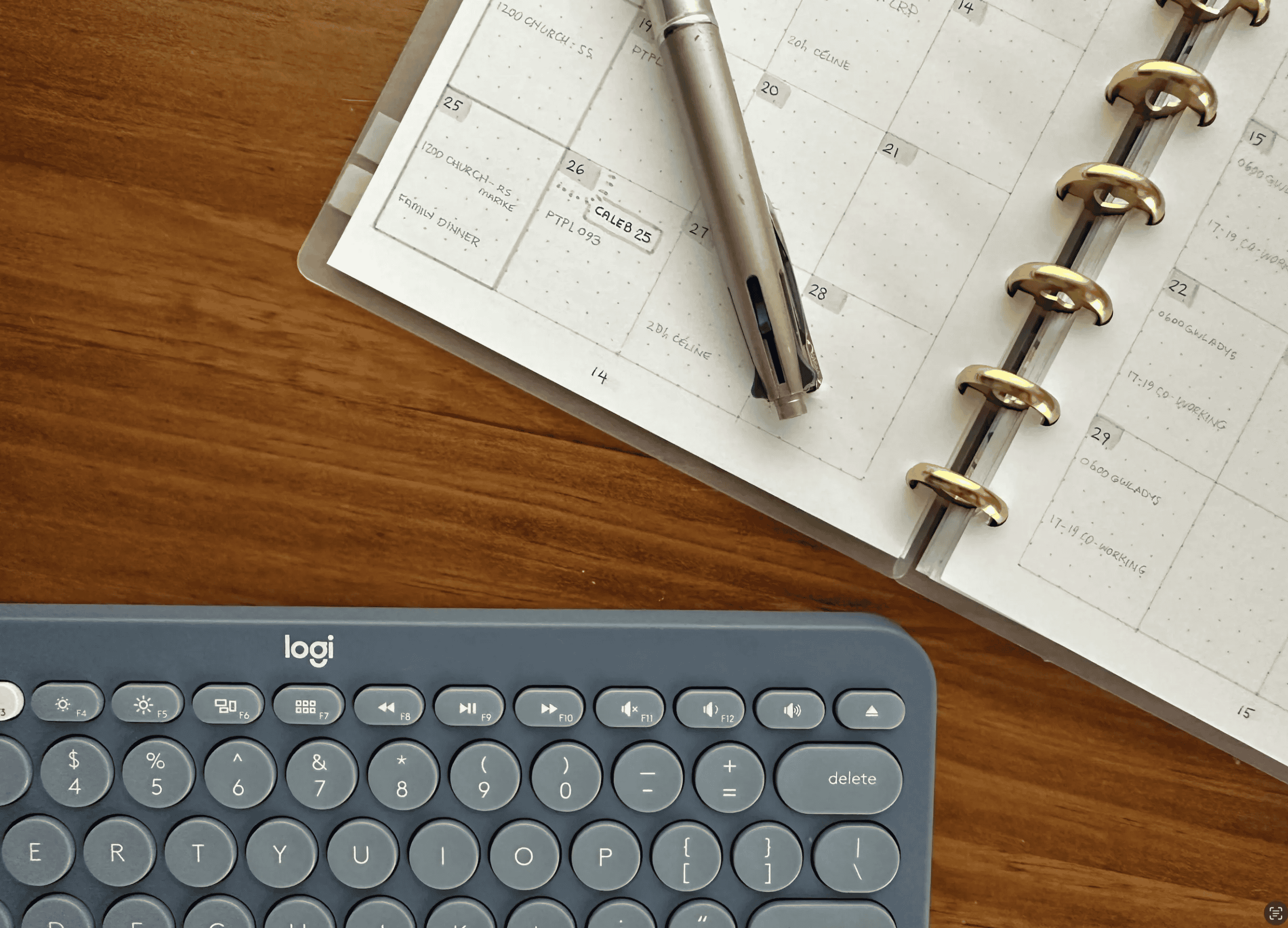 Blue-grey ‘logi’ keyboard and a discbound note book are sitting on a wooden table. We’re looking at a cropped view of them, from above. There’s a Pilot Dr Grip pen (missing its cap) sitting on the left hand planner page.