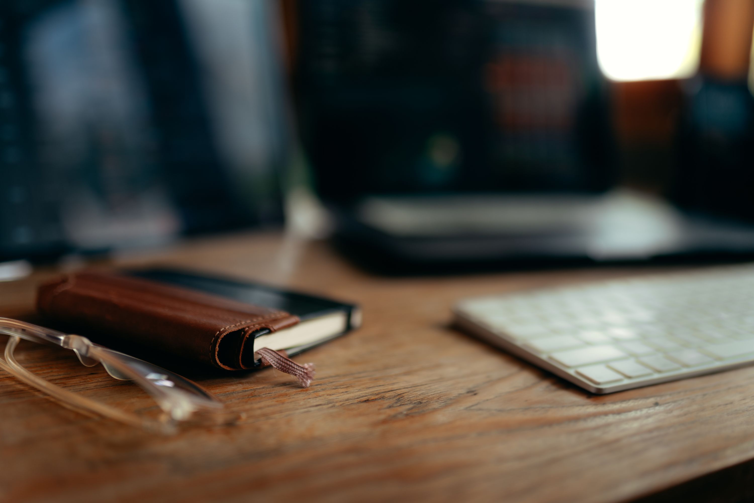 Shallow depth of field photo with partial focus on a small notebook with a leather pen holder on the spine and an external keyboard, with a laptop in the background