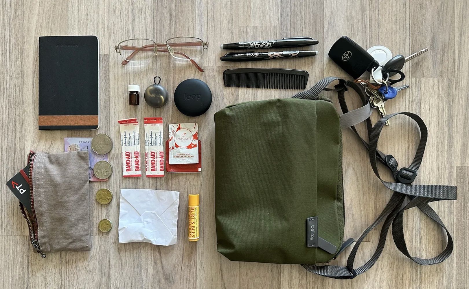 Photo of a khaki green shoulder / crossbody bag next to its contents, which include a notebook, pens, keys, earplugs, bandaids, lip balm, glasses