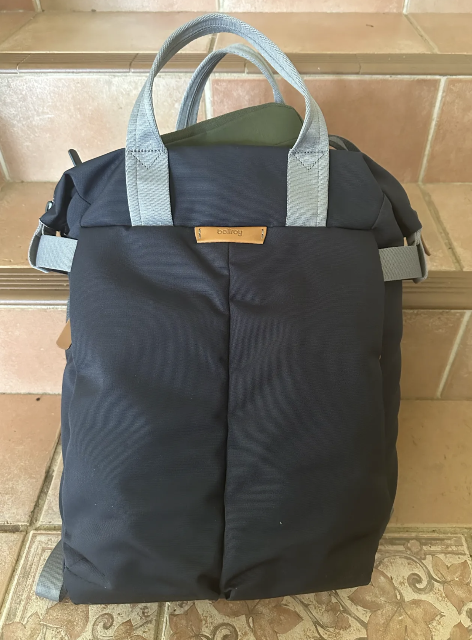 A dark grey bag with a tan leather label is leaning against some steps. The handles are a lighter grey. A tan zipper pull can be seen on the left hand side of the front of the bag, and the author’s khaki cross-body pouch is poking out the top.