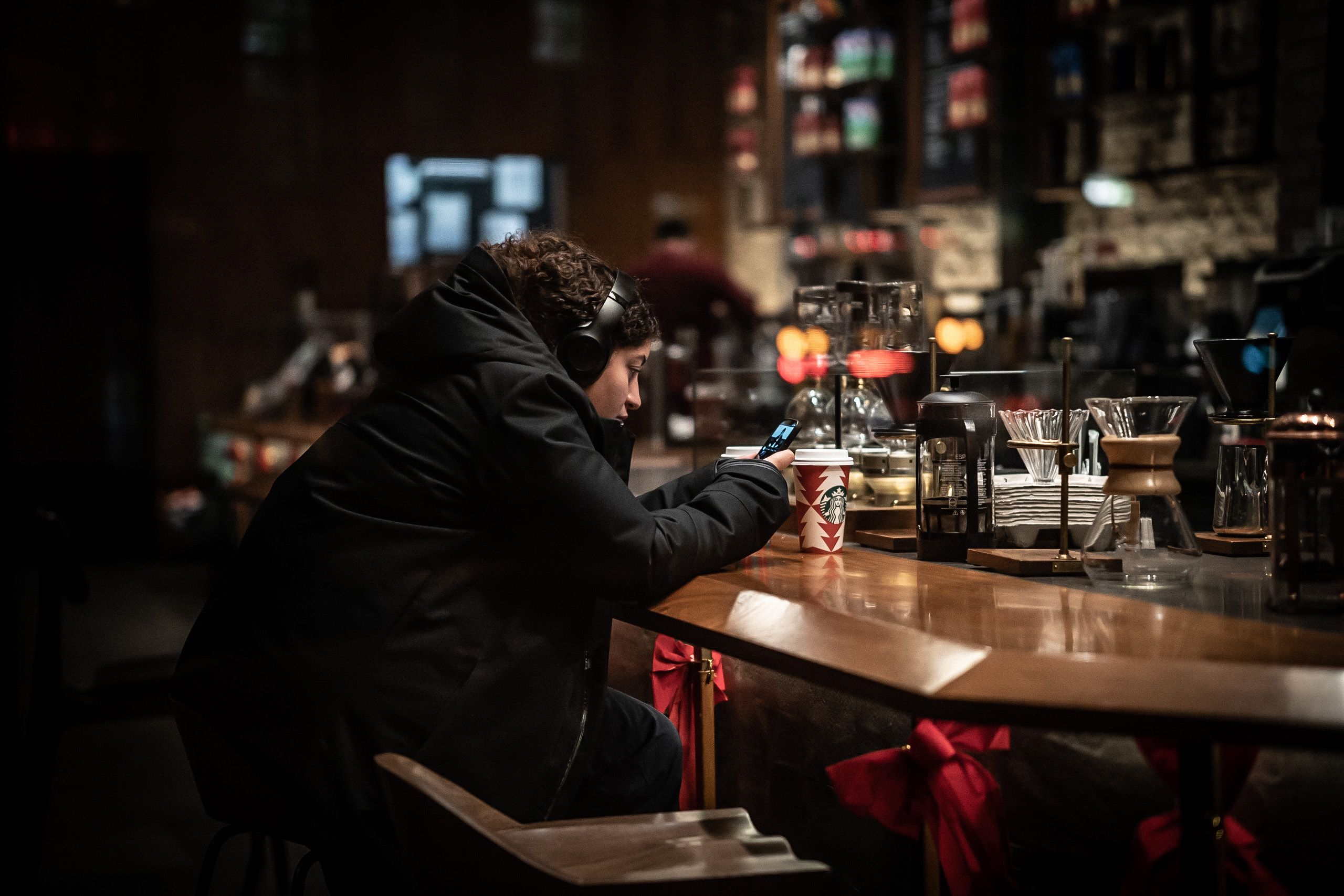 View through a window of a woman sitting at a counter in Starbucks, wearing a winter coat and headphones, looking at her phone