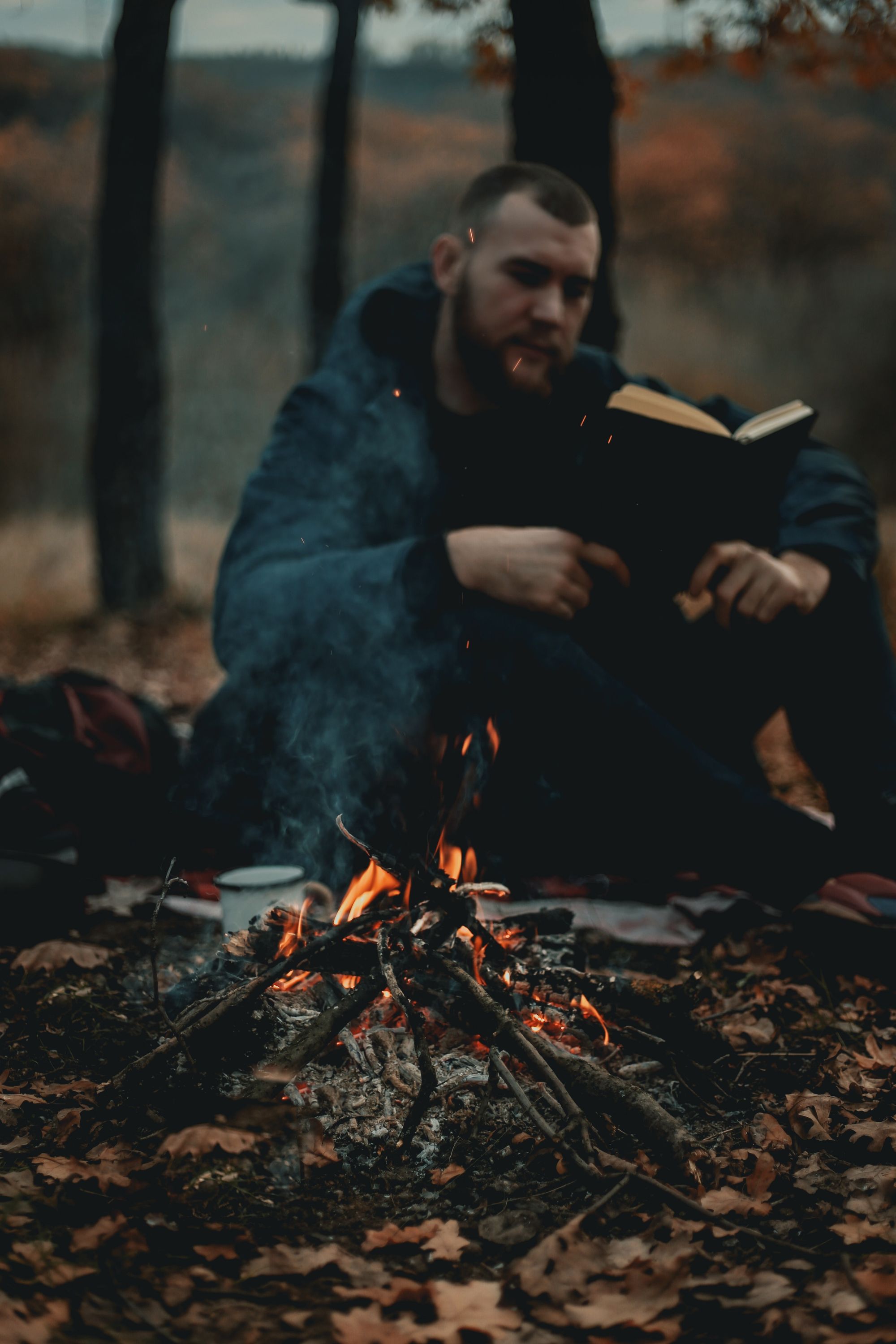 Photo of a man sitting by the fire in the middle of the woods reading.