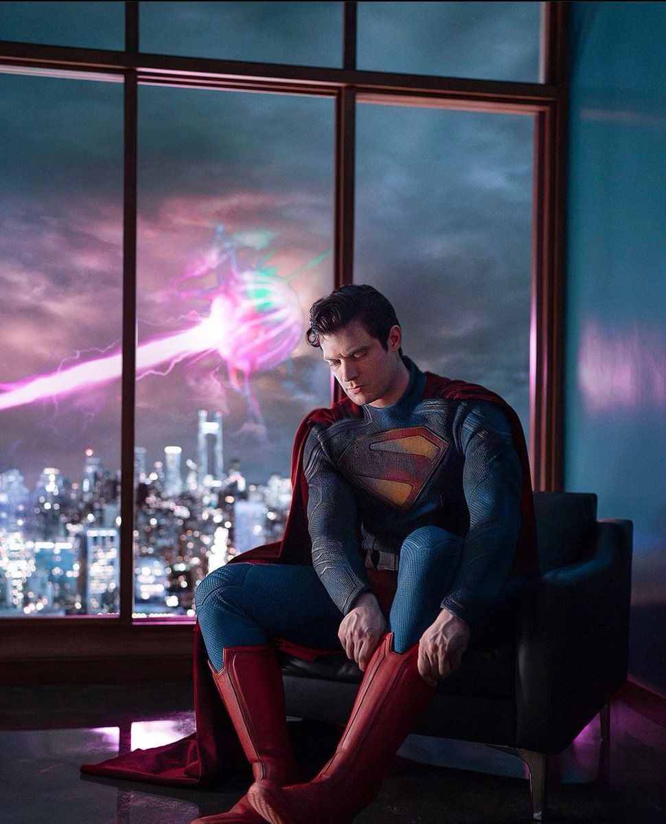 In the foreground Superman is sitting putting on his boots. While outside the window behind him something is firing a pink beam towards the city.