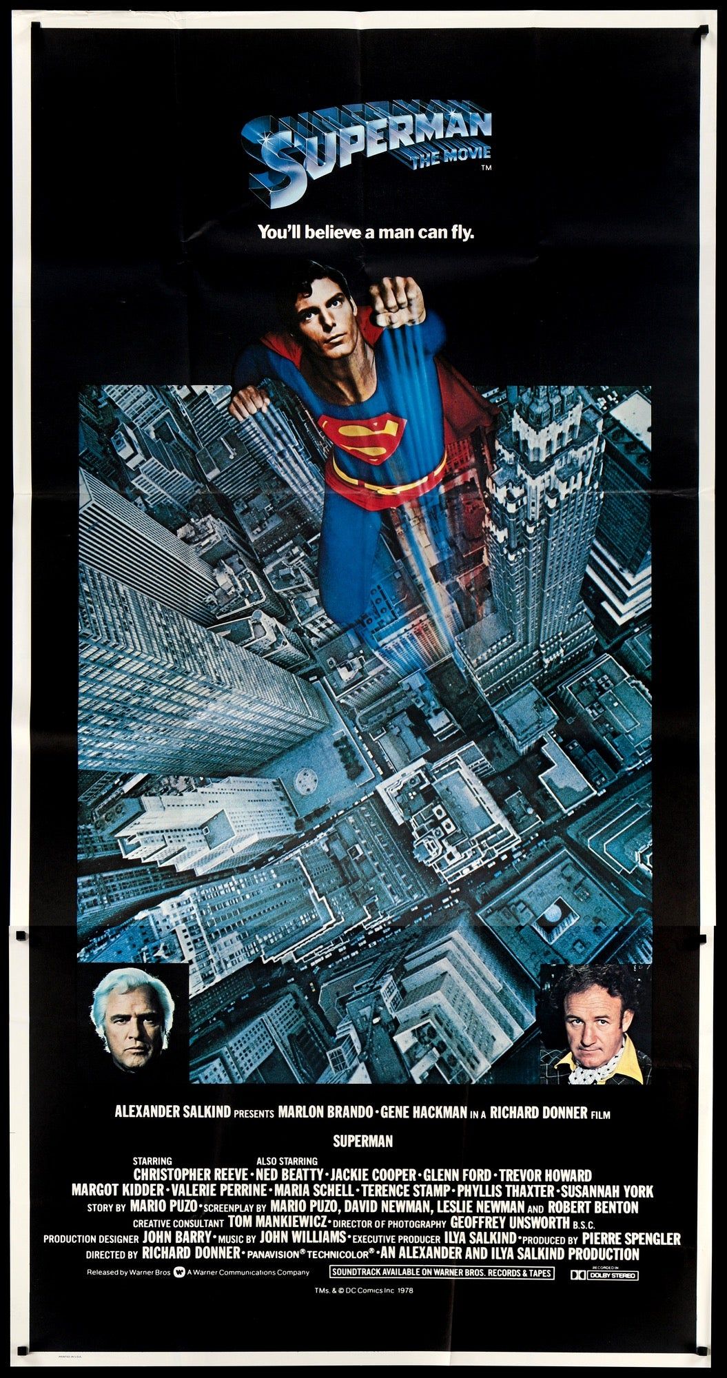 Poster for Superman the Movie with Christopher Reeve as Superman flying up towards the top. Square pictures of Marlon Brando and Gene Hackman in the bottom corners. Text reads Superman the Movie You’ll believe a man can fly.