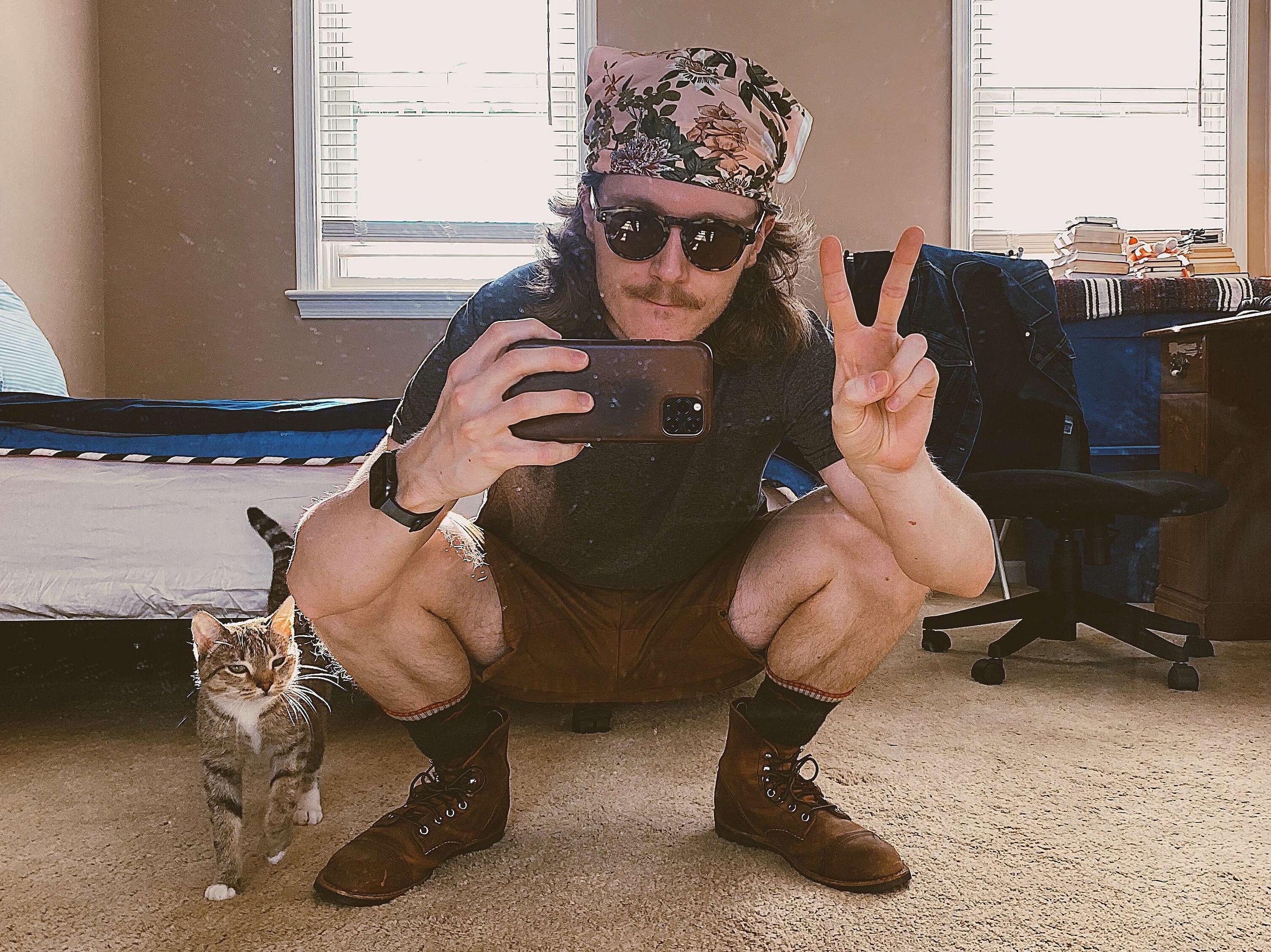 Person squatting in front of a mirror taking a selfie, wearing a floral bandana, sunglasses, and boots, with a cat walking nearby.