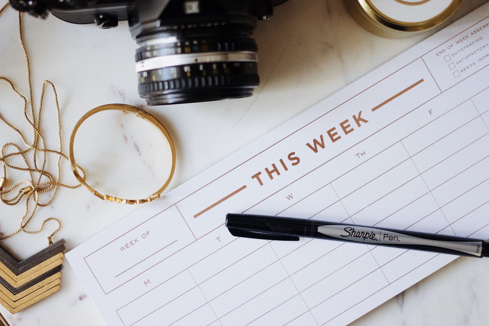 Picture of a blank weekly planner with a Sharpie pen laid across it and some jewellery and a camera around it