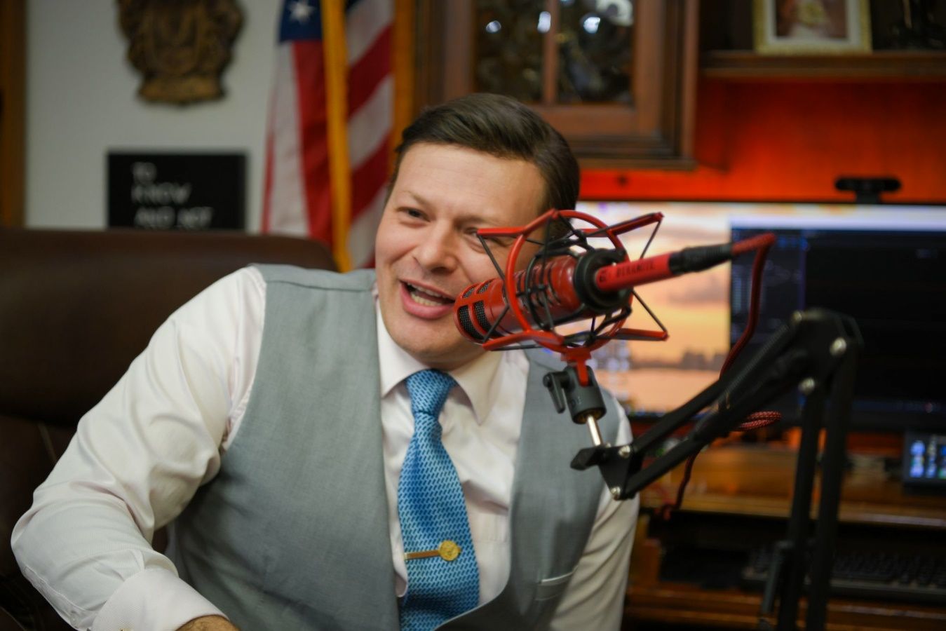 A person in a vest and tie with a microphone in front of him Description automatically generated