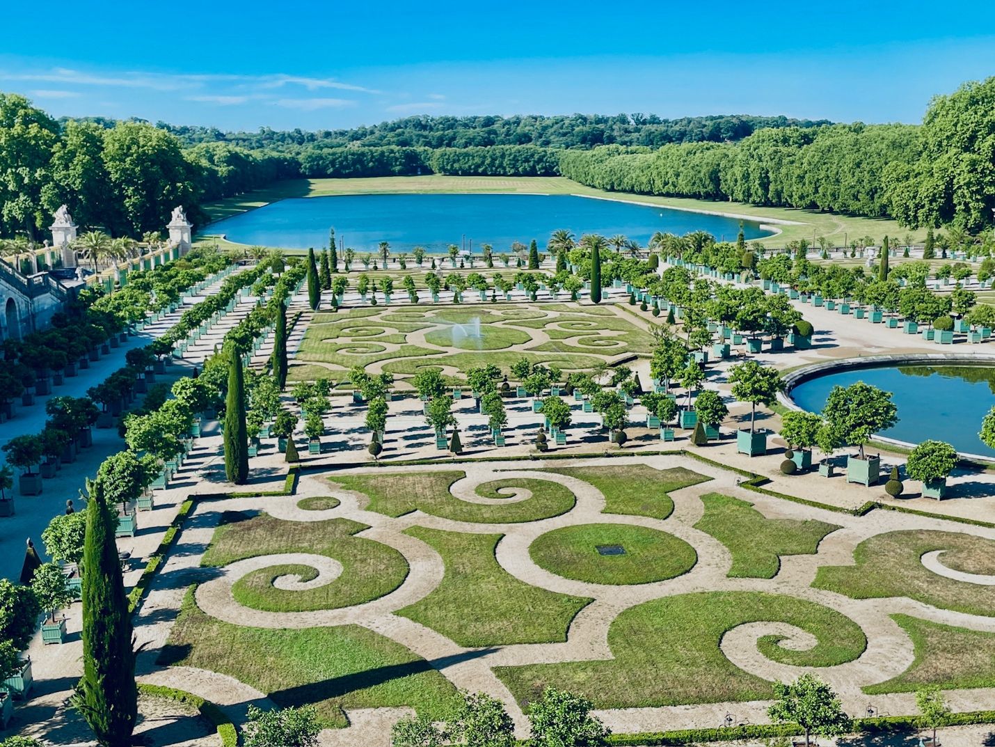 A large garden with a pond and trees with Palace of Versailles in the background AI-generated content may be incorrect.
