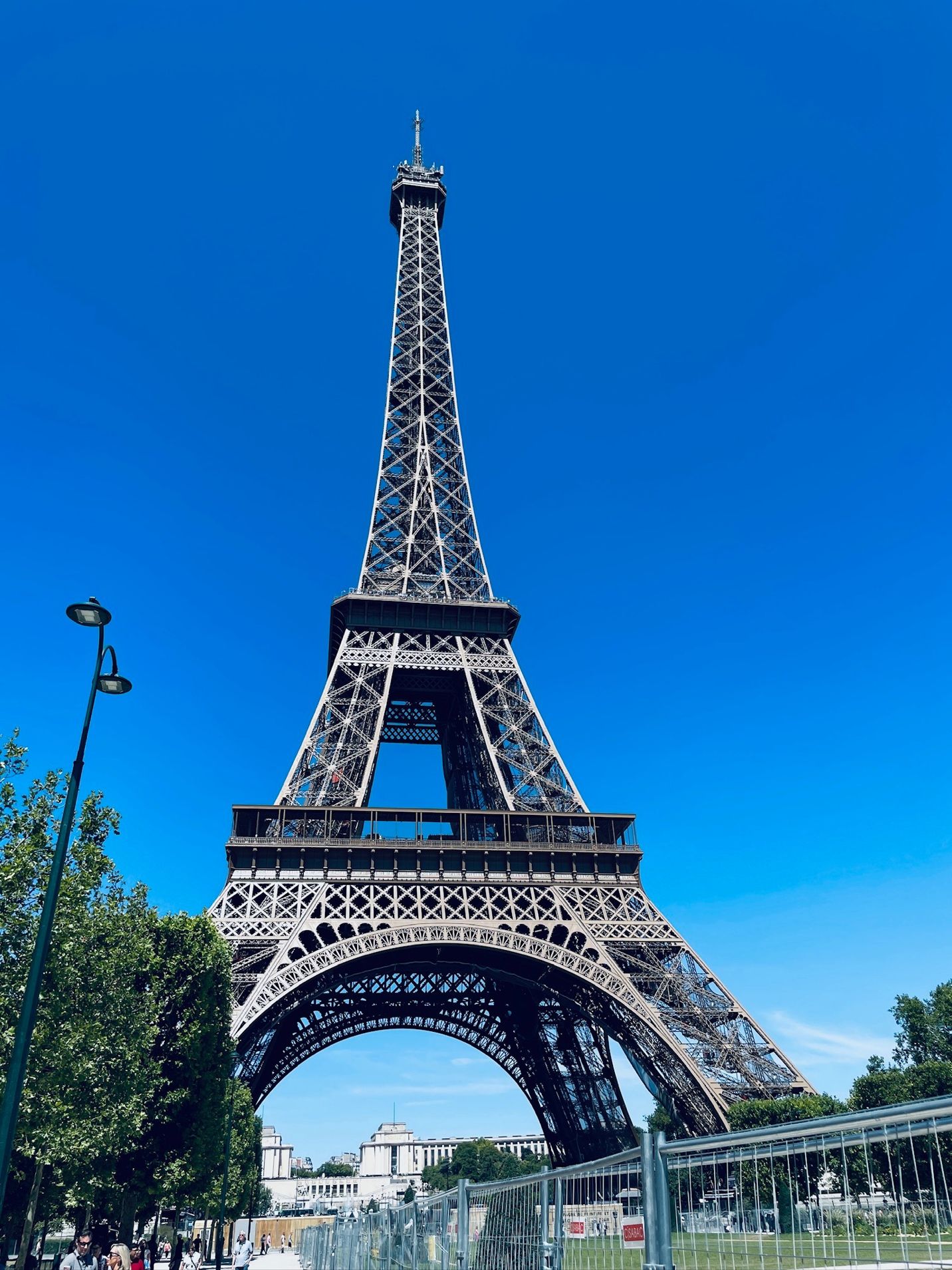 A tall metal tower with trees and blue sky with Eiffel Tower in the background AI-generated content may be incorrect.