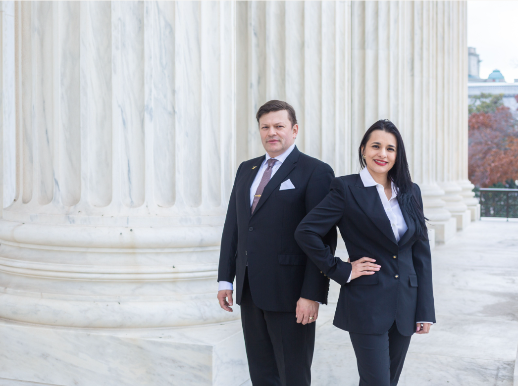 A person and person in suits standing in front of a white column Description automatically generated