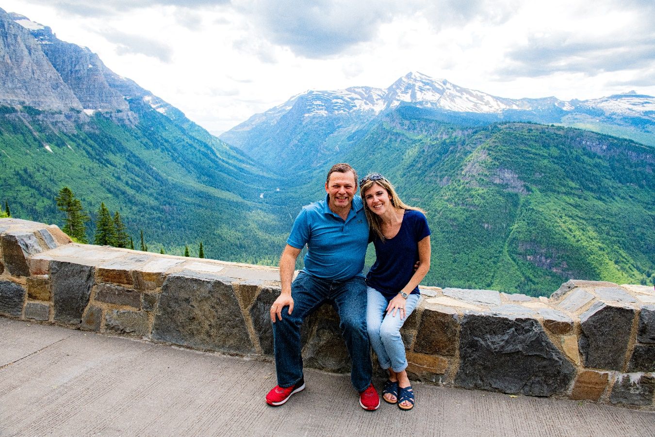 A person and person sitting on a stone ledge with mountains in the background Description automatically generated