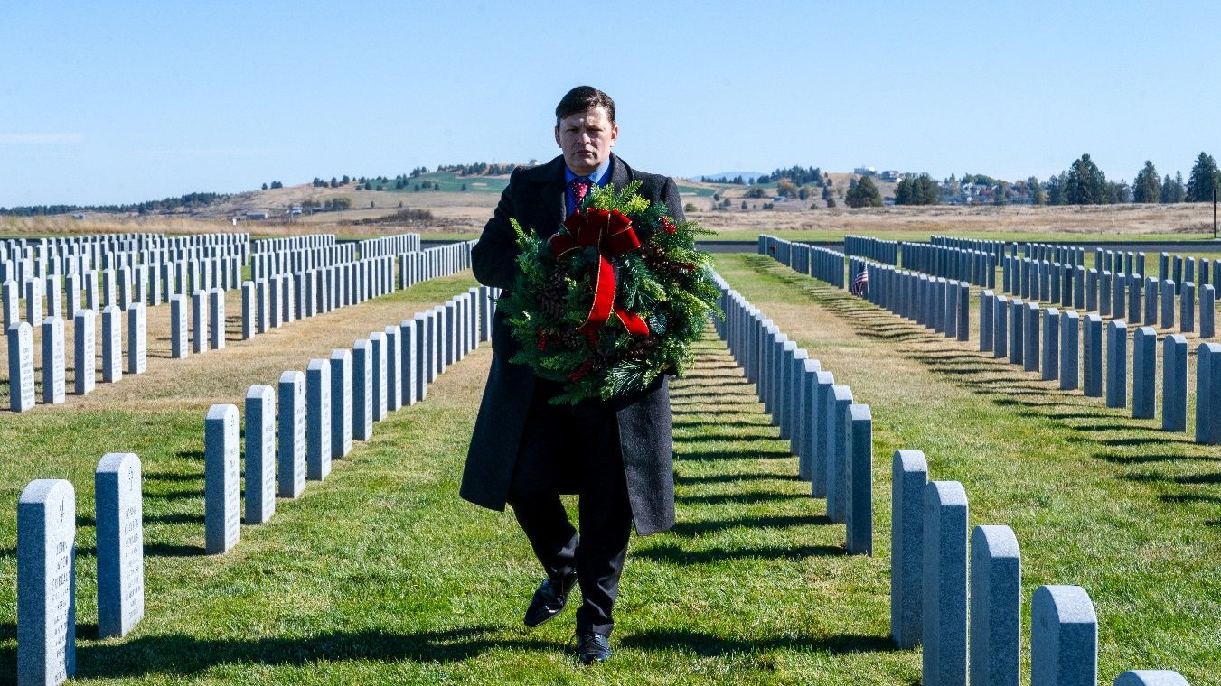 A person in a suit holding a wreath in a cemetery Description automatically generated
