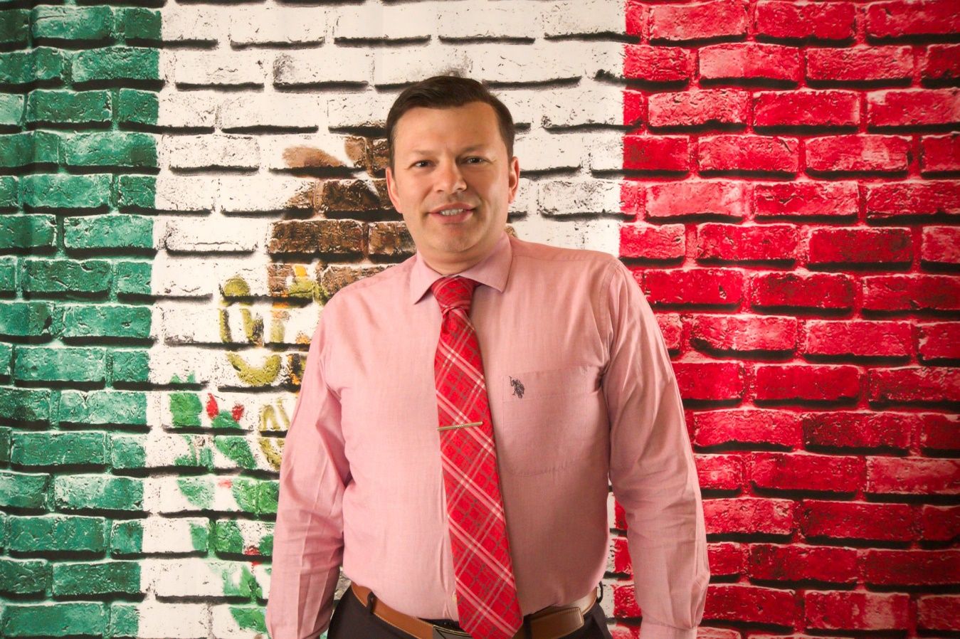 A person in a pink shirt and tie standing in front of a brick wall Description automatically generated