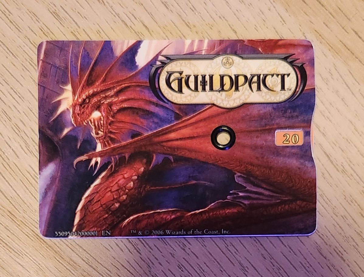 A wallet-sized plastic card with a picture of Niv Mizzet (a dragon) and a “Guildpact” logo. On the right-hand side, an indentation reveals the edge of an orange wheel, and a window inset slightly from that edge reveals a number “20.”