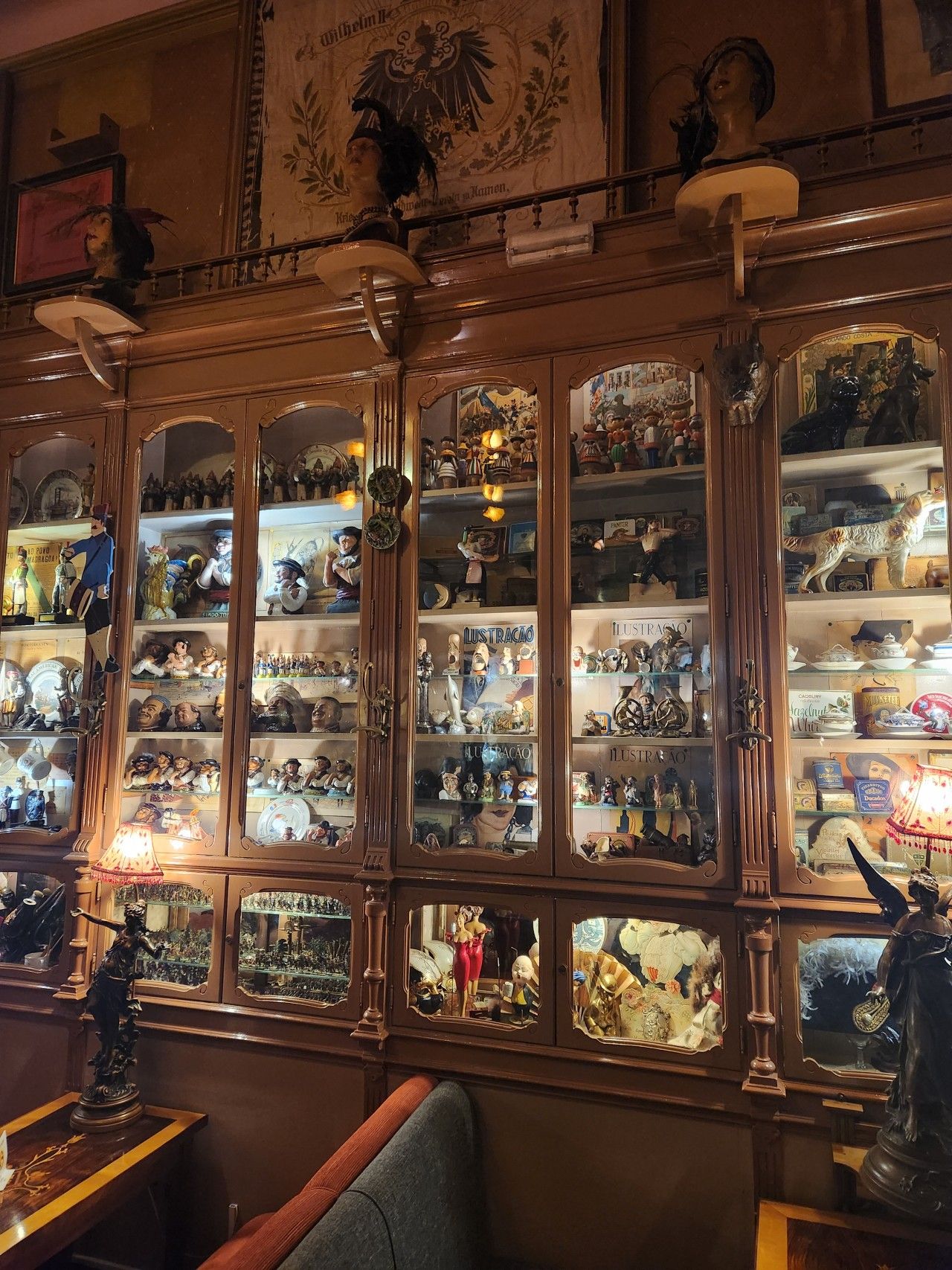 A wall of glass break-front cases filled with figurines of all kinds: armies of miniature soldiers, porcelain dogs, wooden puppets, small lounge singers, teacups, paper ephemera, a slightly-hidden anime figurine, and one case predominantly full of flexing sailors.