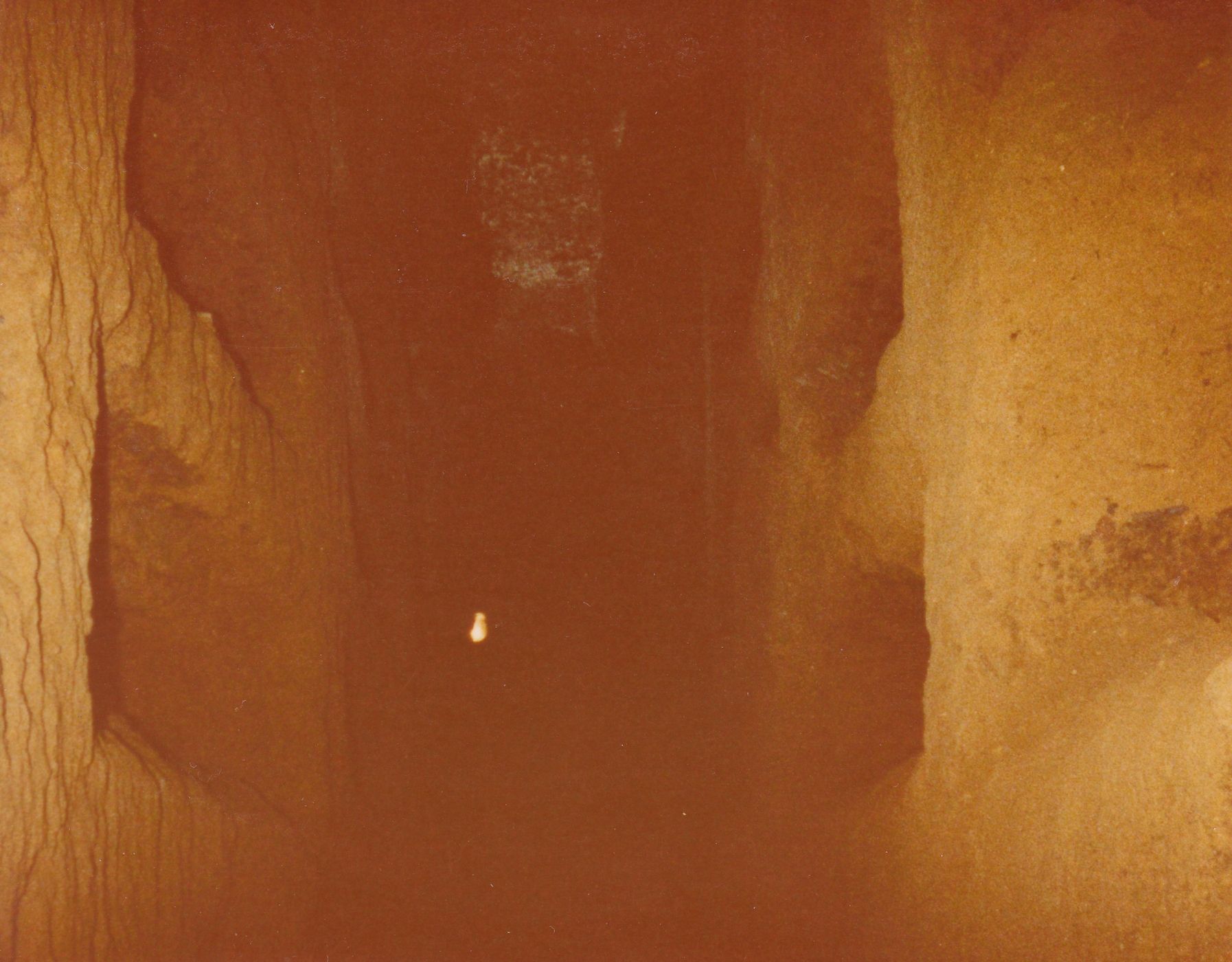 A washed-out photograph of a narrow hewn-stone hallway with long niches carved into the walls. There is a light source in the distance, but surrounded by darkness.