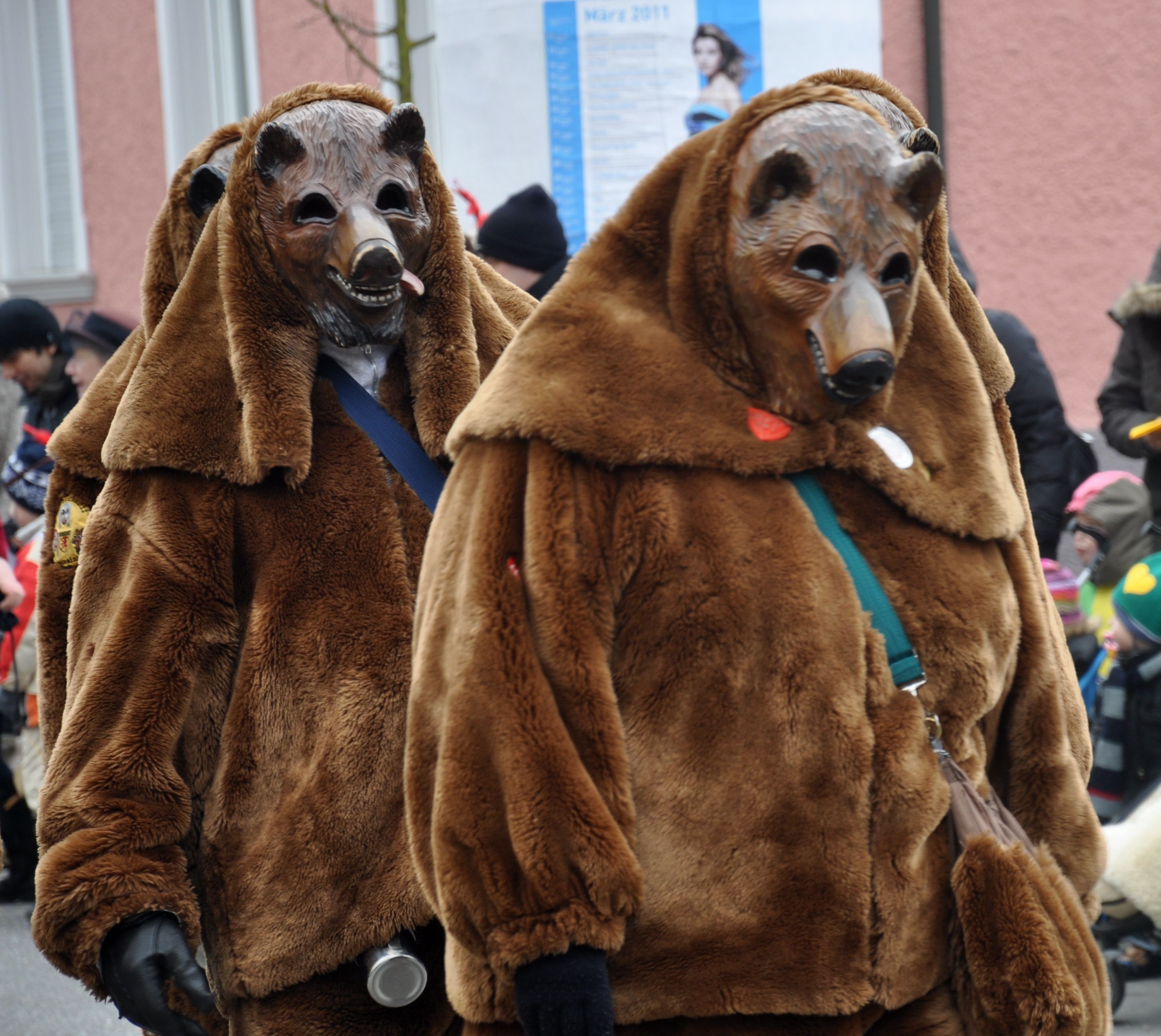 A photograph of a procession of figures (at least three) in brown bear costumes with colorful buttons and cross-shoulder bags.