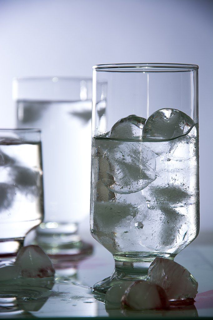 A short-stemmed drinking glass full of round ice cubes and water. Two other glasses are behind it in the background, and some ice cubes are melting outside of the glasses on the table.