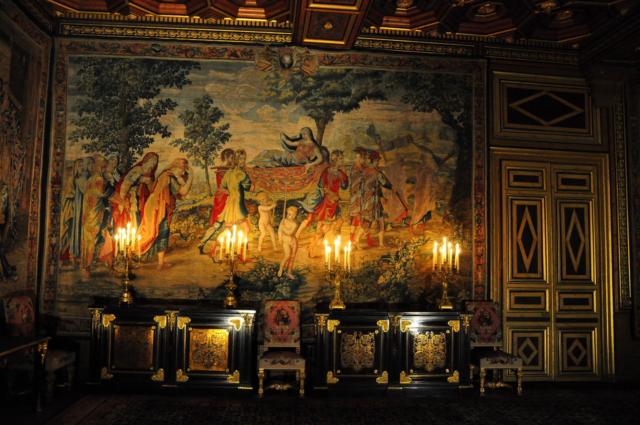 A medieval tapestry in a darkened ornate room. The only light comes from four candelabras in front of it, catching the gilt surfaces of all the framing of the room and furniture.