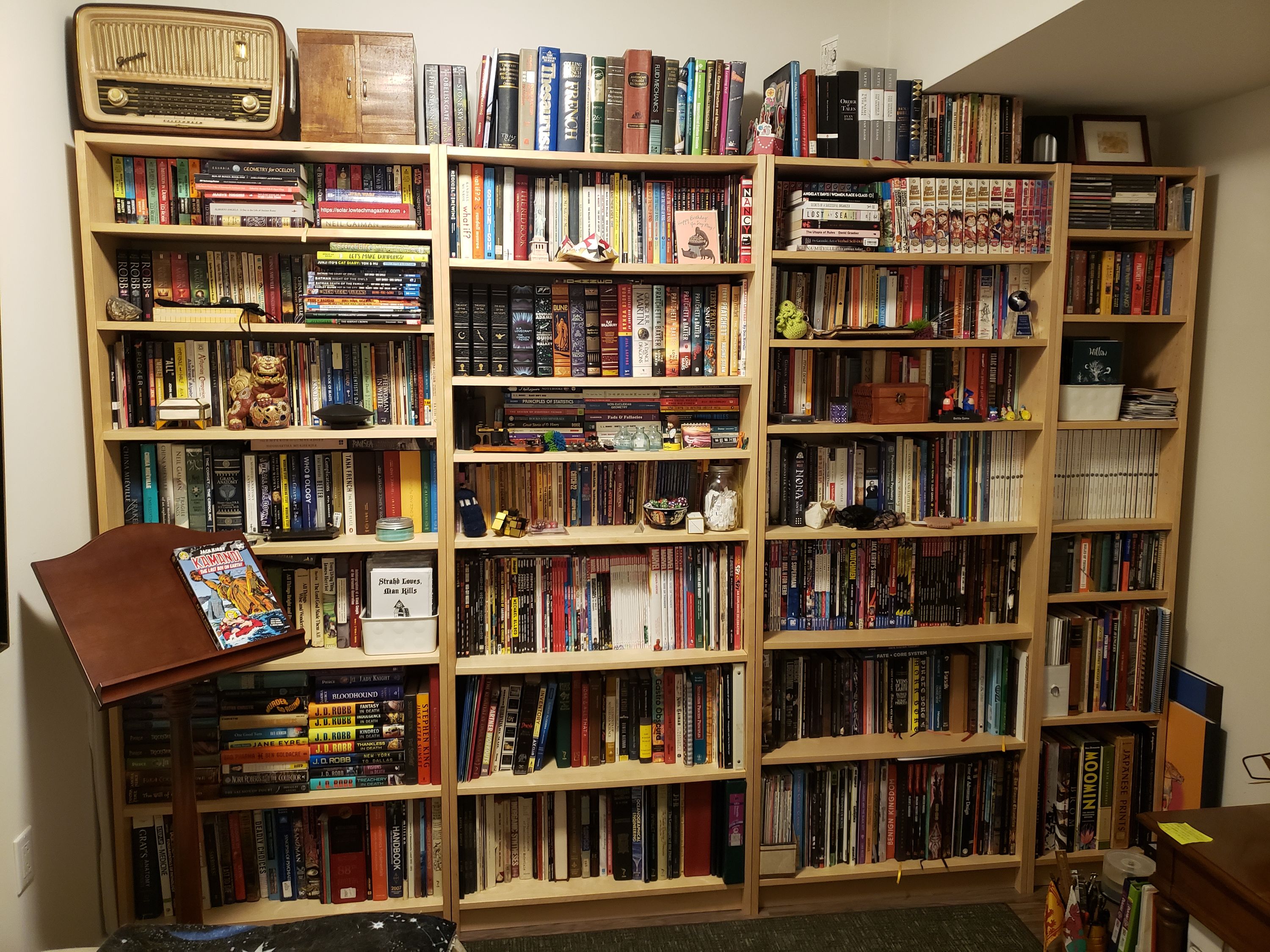 Three and a half full-height bookshelves are anchored against a wall, practically filling it. There are too many books on them and on top of them, stacked in no reasonable way, complete with more tchotchkies.