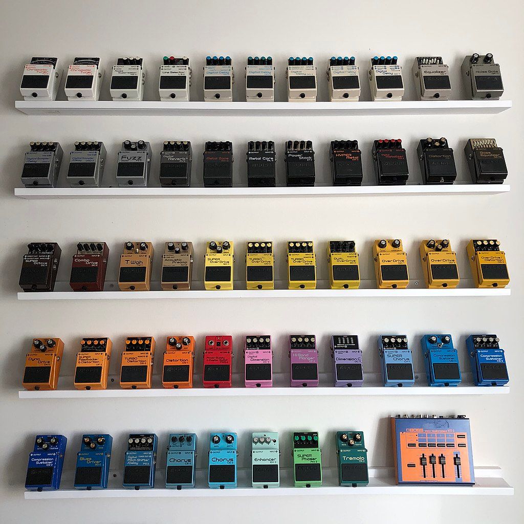 Several dozen guitar pedals arranged by color across five shelves.