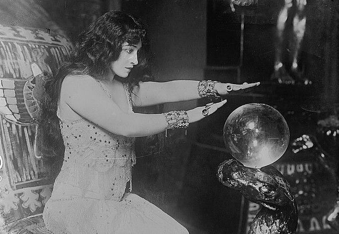 A black and white photograph of actress Pauline Frederick, seated and gazing into a crystal ball with her hands outstretched.