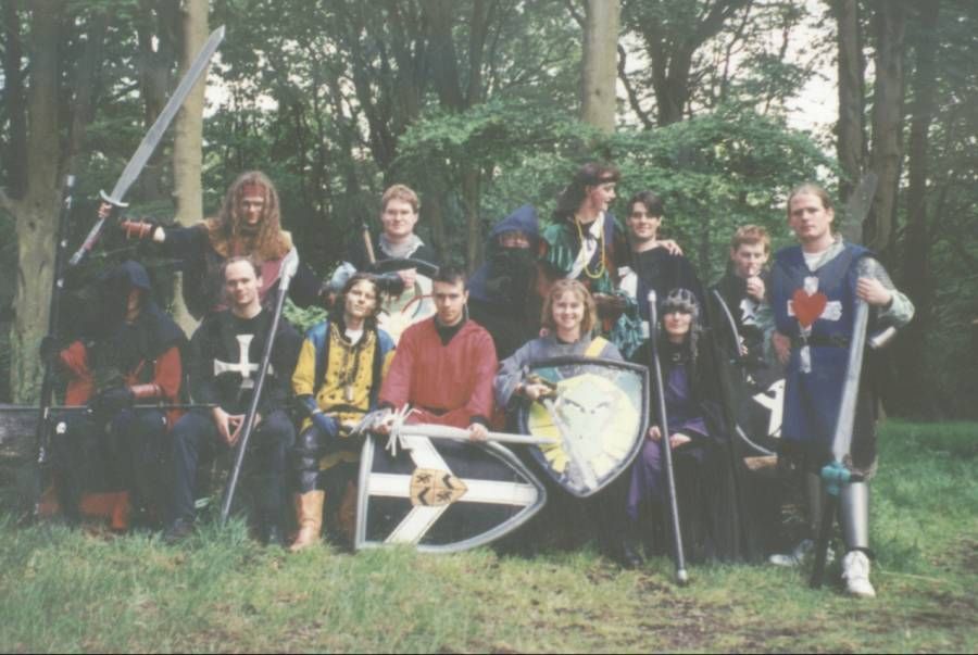 A photograph of 13 Medieval LARPers in the woods from Birmingham University in 1997. They pose for the camera with armor, shields, and swords.