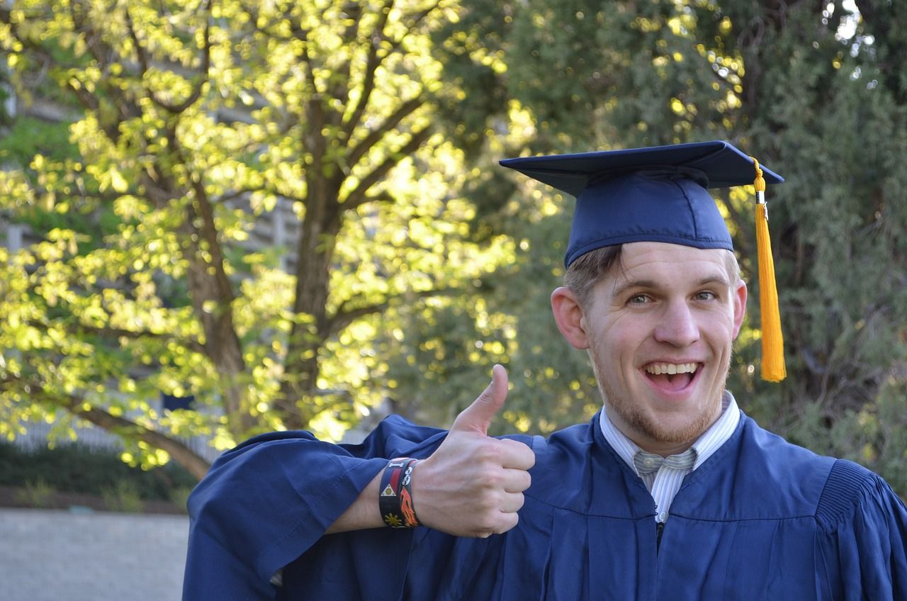 A grinning guy in a blue mortarboard-hat and robes gives an exaggerated thumbs-up to the camera, in front of some trees.
