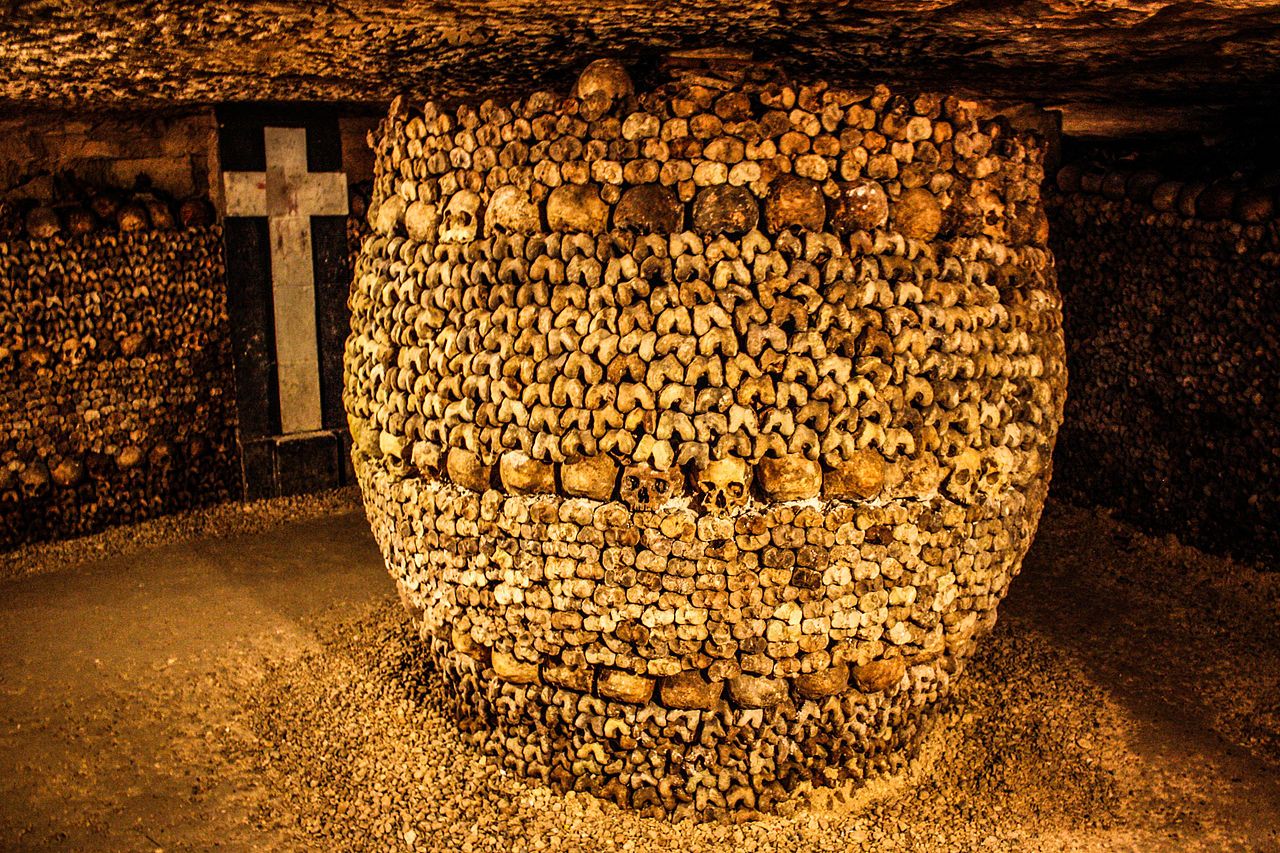 A wide pillar of bones with organized rows of human skulls dominates the center of a dark, low-ceilinged room. A large cross if visible set into the wall behind it.