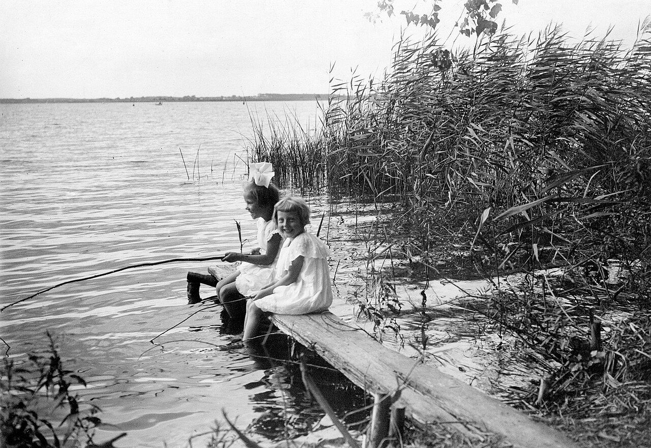 A black and white photo of two young German girls seated on a board, fishing with sticks in a wetland. The closer one grins at the camera.