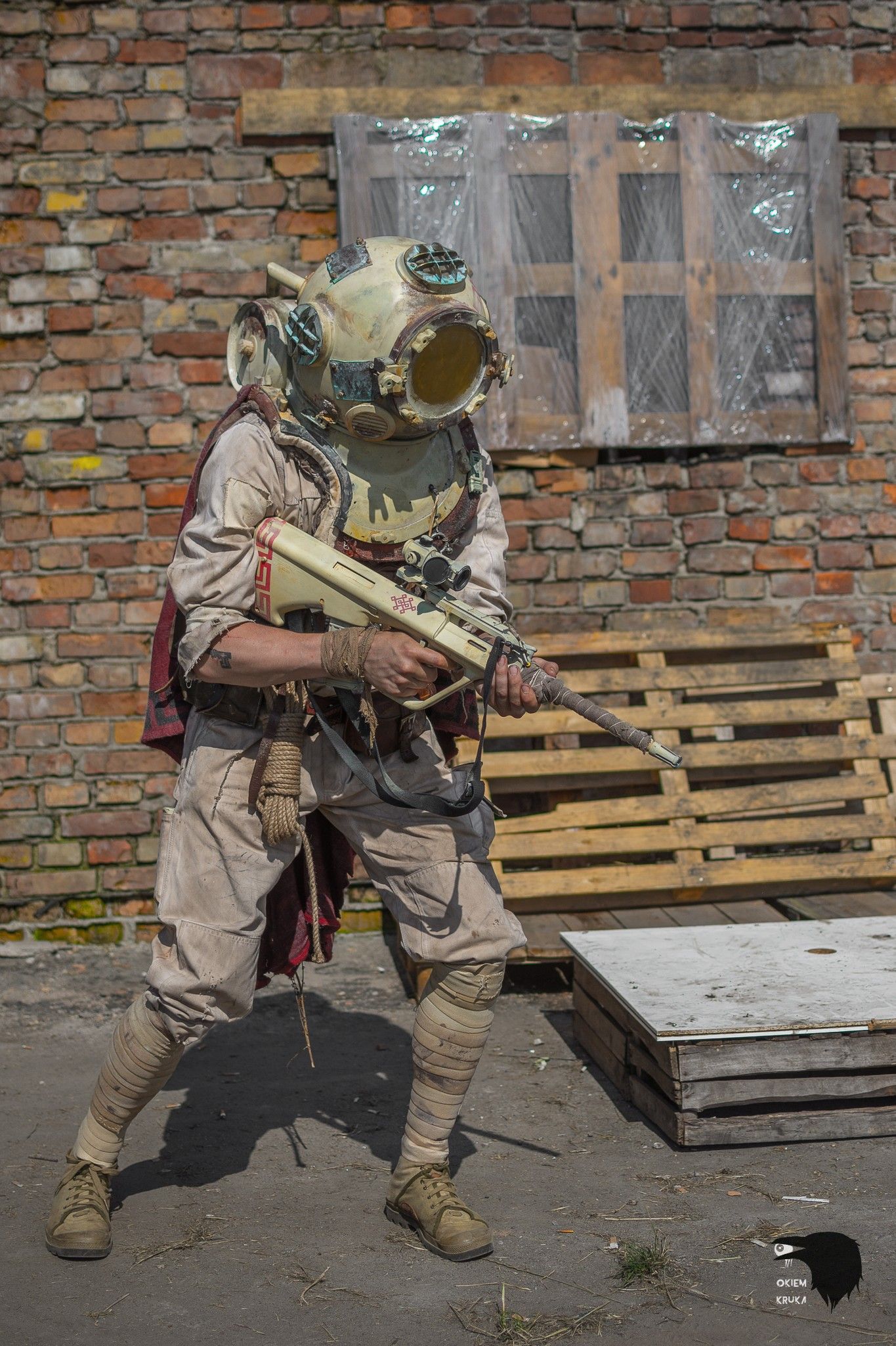 A diving-helmeted soldier carries their rifle in front of a brick wall with some pallets.