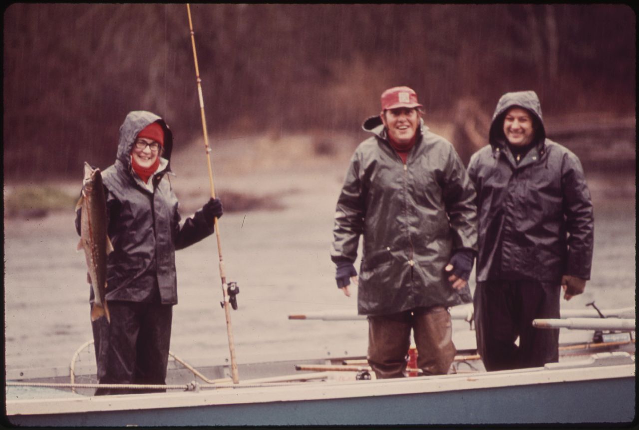 A blurry photograph of three people in a boat. A grinning woman on the left holds a large trout and a fishing rod. To her left, two men also smile for the camera. All three are dressed in heavy raincoats.