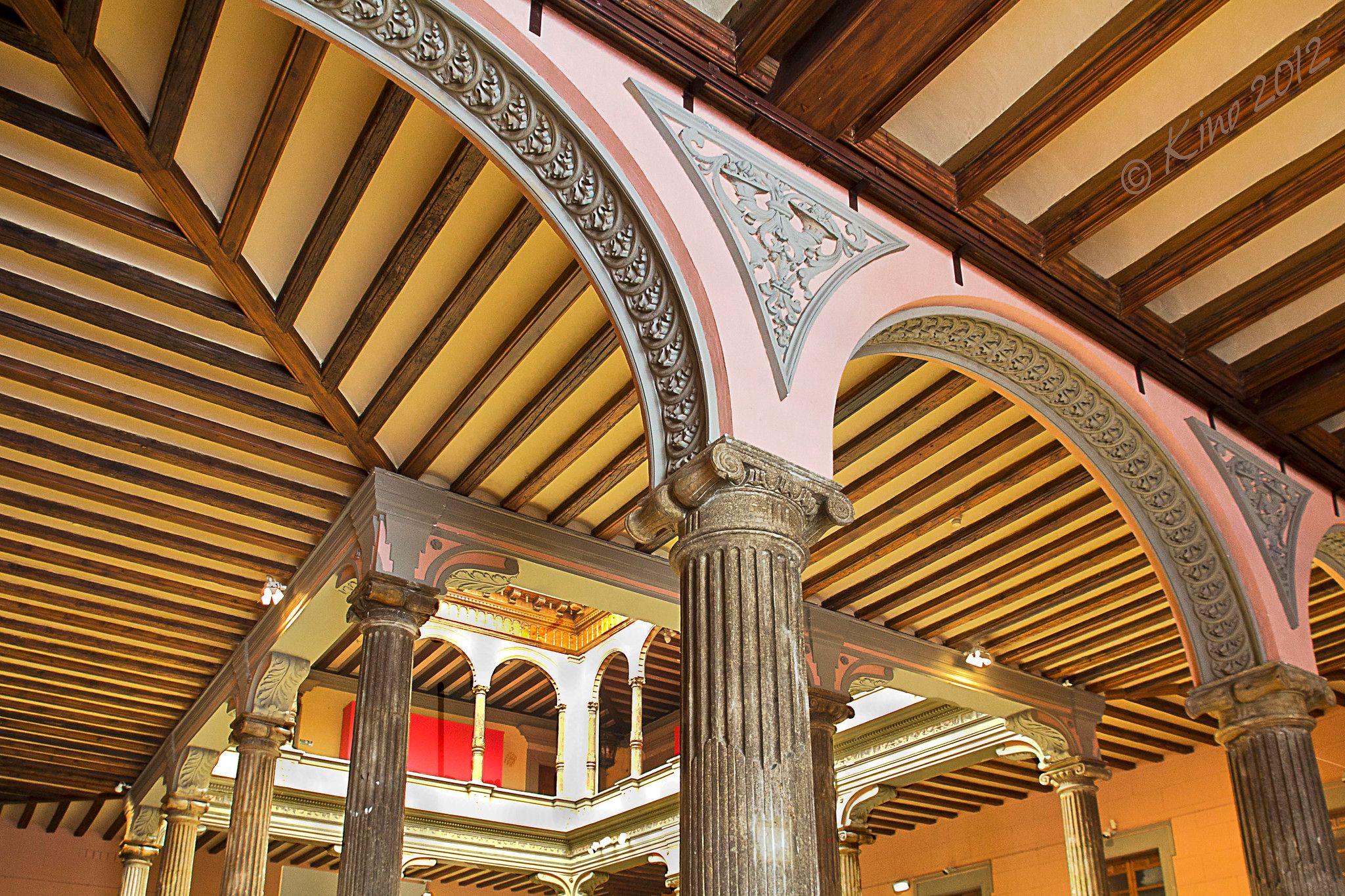 The ceiling of a palace, flat with many small beams, held aloft by columns and arches. The sun filters in through a large square opening, through which can also be seen the arches of a second story.