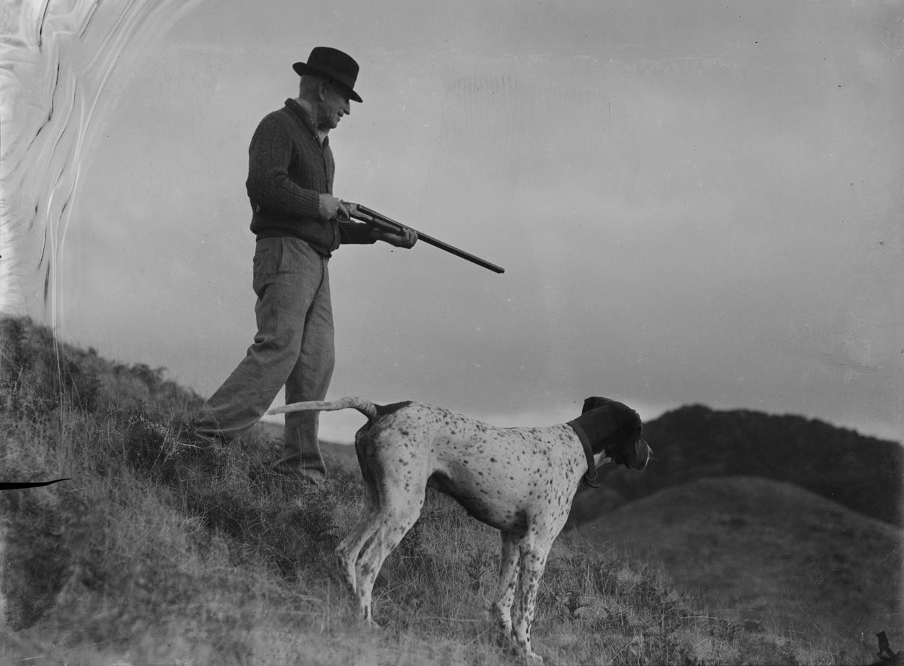 A slightly damaged black and white photograph of a man in modern dress holding a hunting rifle and with a dog, in some hilly landscape.