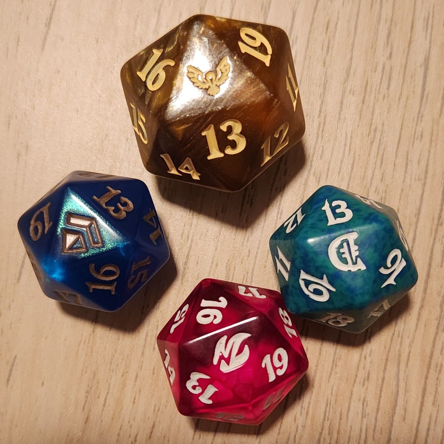 Four “spindown” style d20 life counters. The face-up symbols on each are from Strixhaven, Amonkhet, Deckmasters, and Fire & Lightning. The Strixhaven die is larger than the others. Surrounding the symbols replacing “20” on each are sides with 19, 16, and 13.