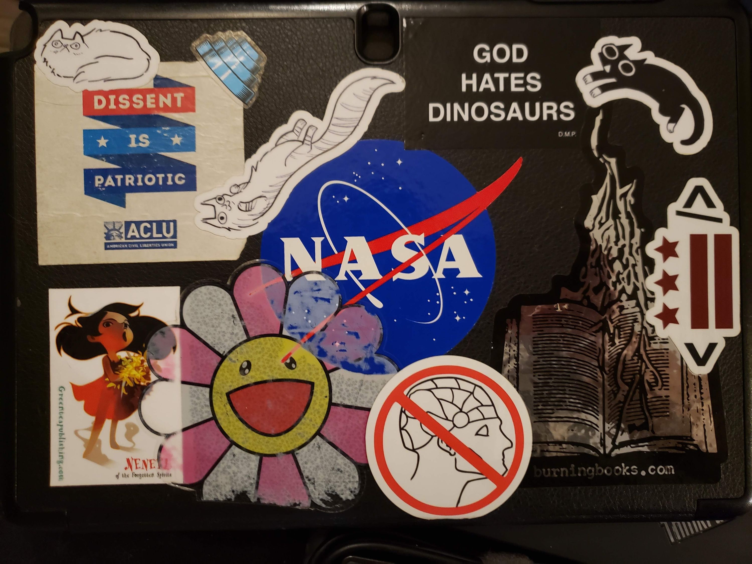 An empty tablet case, covered in stickers. “Dissent is Patriotic” from the ACLU, a NASA “meatball” logo, and a shiny “burningbooks.com” logo largest, followed by a faded and worn smiling Murakami flower, a small girl (Nenetl of the Forgotten Spirits), a phrenology head diagram with a cancel sign through it, A DC flag enclosed in angle brackets like an HTML tag, and a clean “GOD HATES DINOSAURS.” several cartoon cats and a Devo energy cone decorate the smaller spaces.