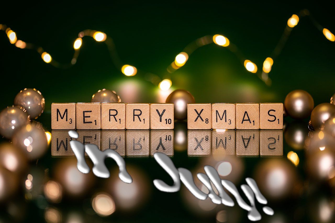 Scrabble tiles spell out “MERRY XMAS” on a mirrored surface, surrounded by out-of-focus lights and baubles. “in July!” appears superimposed in a “fun” font with a drop-shadow.