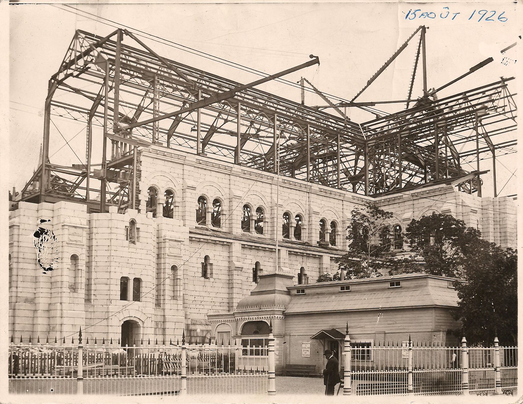 A sepia-toned photograph of a cathedral with walls half-built and cranes above it. A man in a suit breaks the foreground. Captioned in blue pen: “15 Août 1926”.