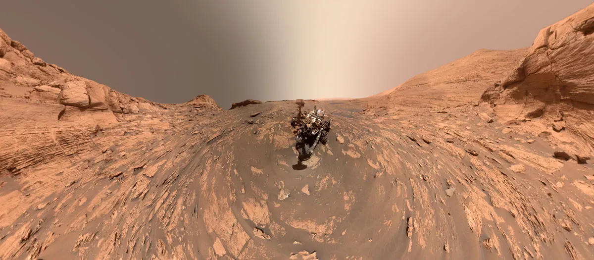 A 360-degree panoramic martian landscape, dusty red and brown with an unremarkable grey sky. In the center, the Curiosity rover.