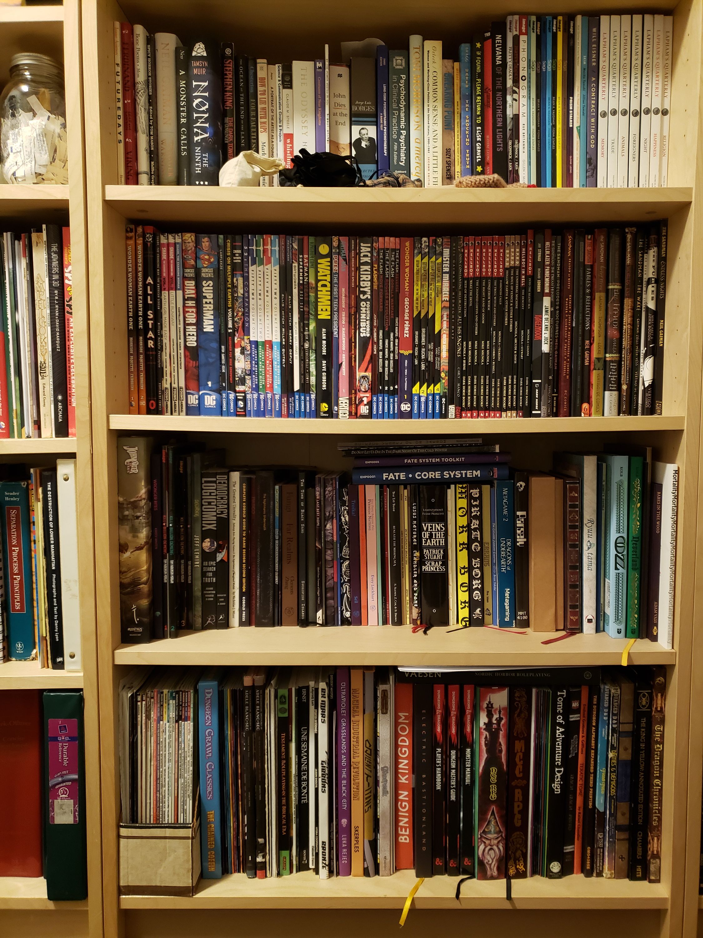 A close-up of the bottom four shelves of the third-from-the-right case in the earlier photograph. The top shelf is miscellaneous, the shelf below it is graphic novels, and the two bottom shelves are mostly RPG books, with the taller books on the bottom shelf.