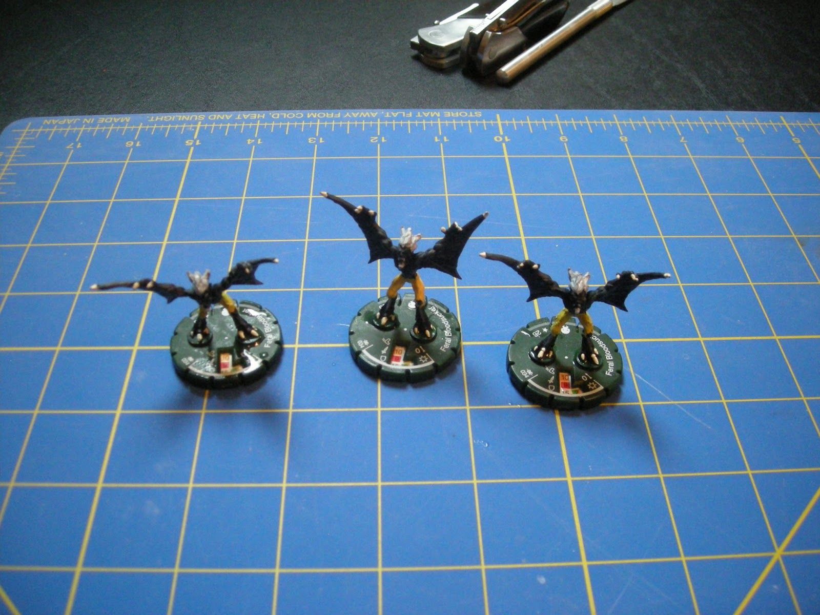 Three black bat-winged Mage Knight figures with white claws and hair and yellow pants stand on a blue and yellow cutting mat.