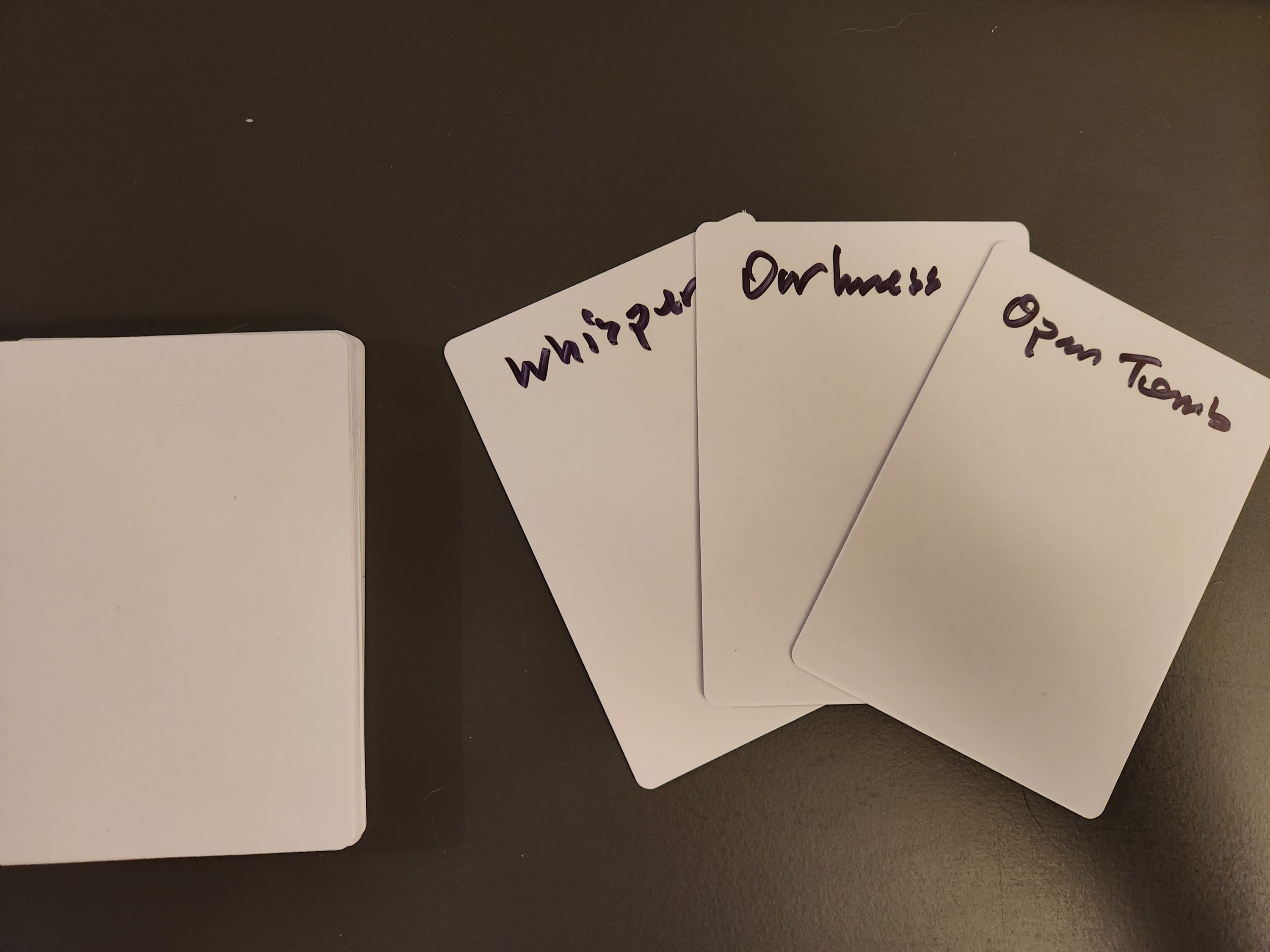 A small stack of blank playing cards. Three face-up cards read “Whispers,” “Darkness,” and “Open Tomb.”